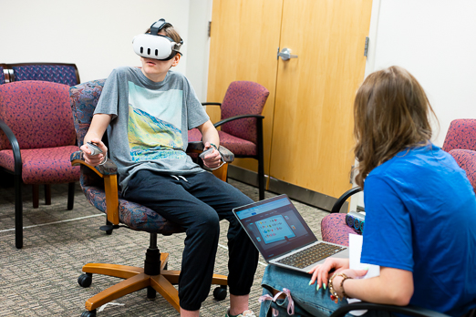 a girl sitting with a virtual reality headset while she's observed by a woman with a laptop