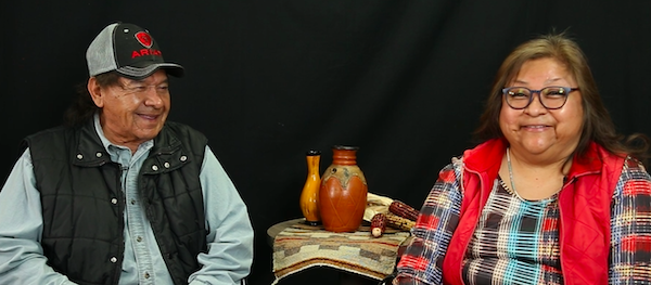 Native American man wearing a black cap and black vest on the left and a Native American woman wearing a bright plaid top with a red vest on the right, both smiling, with artifacts on a small table between them.