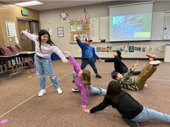 Group of 6 students all working together to make lines, shapes, and levels with their bodies, some lying down, some with arms outstretched, some reaching from the ground to the sky.