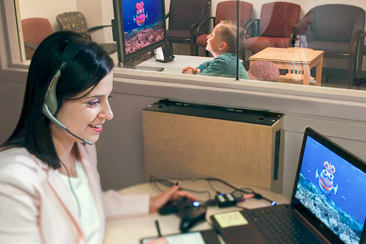 Woman with a headset and computer with a fish on the screen while a boy is on the other side fo the window looking at the same fish