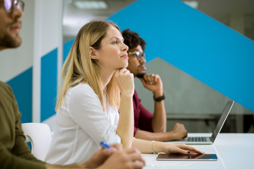 3 people at a desk with their laptops, paying attention to someone speaking out of frame.