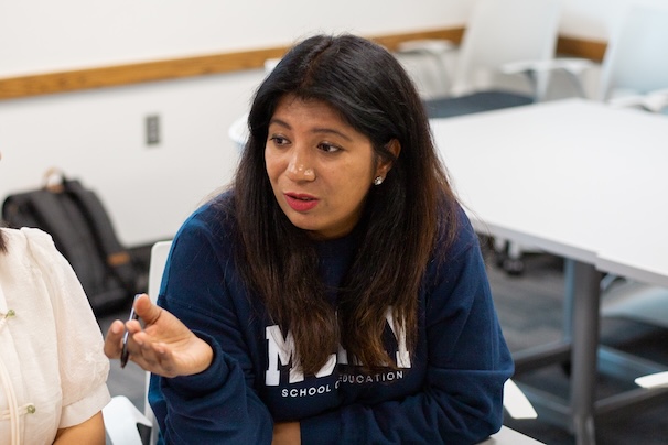 A woman wearing a blue sweatshirt with 'School of Education' written engaged in a discussion in a classroom setting