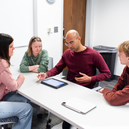 two men and two women at a long table while one man explains something on a tablet