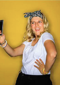 A girl holding a phone up for a selfie who is standing in front of a yellow wall